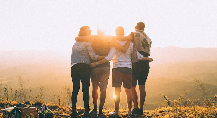 Four friends standing close together, arms around each other, watching a sunset over a scenic landscape.